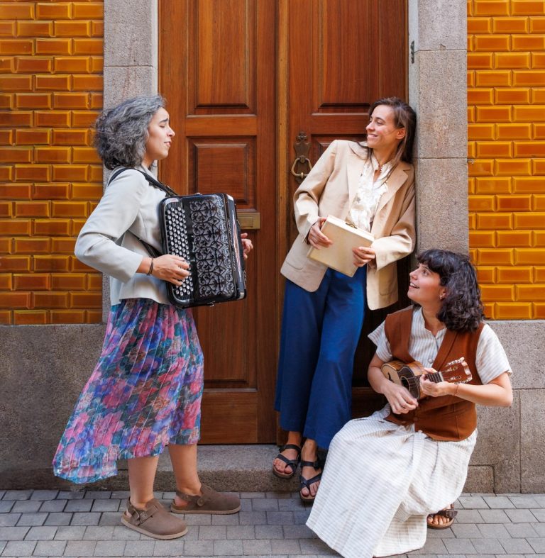 Three women playing music on a sidewalk outside a building: a woman with an accordion stands left, another holds a folder and smiles near the door, and a third sits on the step playing a ukulele, against a yellow brick wall.
