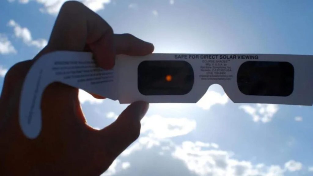 Holding up solar viewing glasses to the sun, showing the orange filter window and the 'SAFE FOR DIRECT SOLAR VIEWING' label.