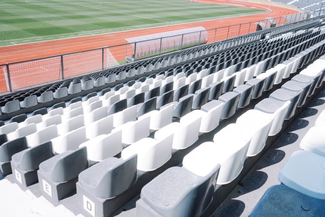 Empty stadium seating with alternating gray and white seats overlooking a green field and red track edge of the stands.