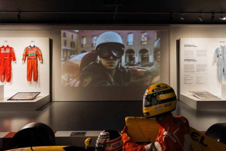 Museum display: orange racing suits on the left, a video of a helmeted driver in a car on the screen, and a yellow race car in the foreground.
