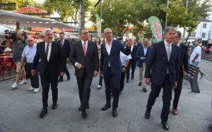 Group of formally dressed men walking together through a city square, with market stalls and onlookers in the background, one man gesturing with his hand.