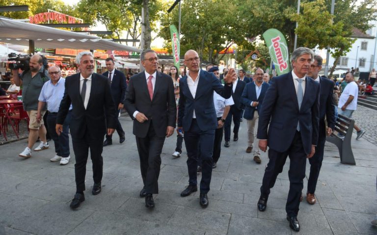 Group of formally dressed men walking together through a city square, with market stalls and onlookers in the background, one man gesturing with his hand.