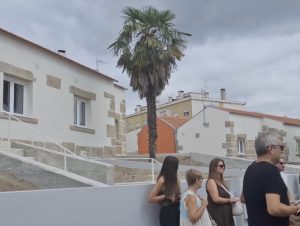 Group of adults standing and chatting near a row of white houses with stone accents and a tall palm tree, on an overcast day.
