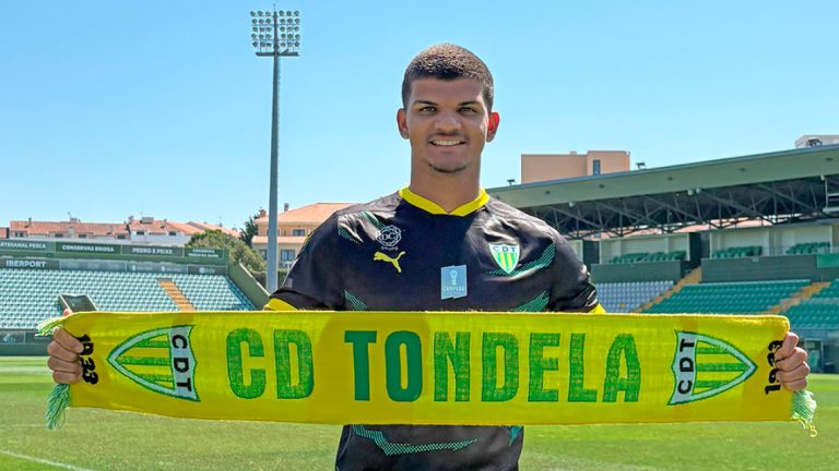 Young soccer player in a black and green kit holds a yellow CD Tondela scarf on the pitch of a sunny stadium.