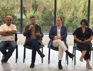 Four people seated in a panel discussion: the second person speaks into a handheld microphone while the others listen; large windows with greenery behind.