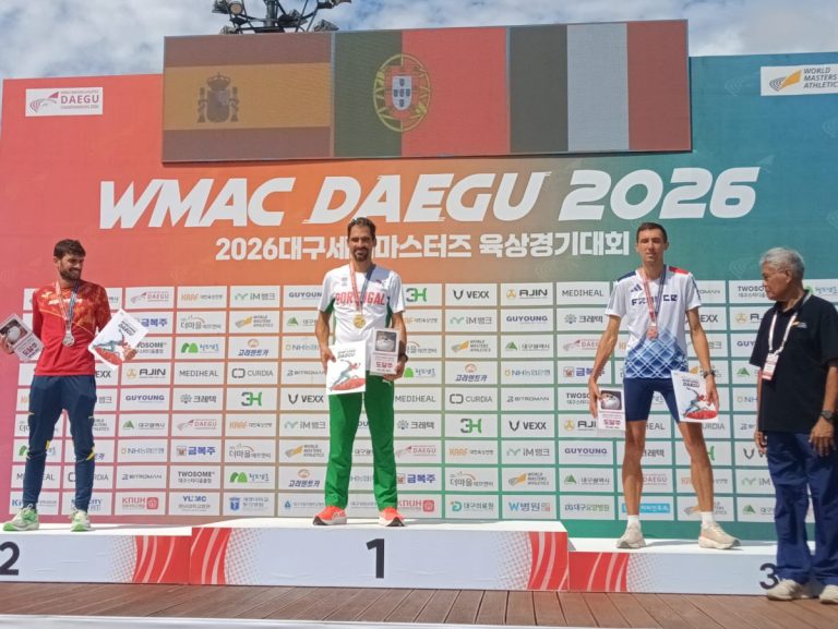 Athletes stand on a podium: first place in center with green pants and white shirt, holding a certificate and wearing gold medal, flanked by second and third place winners on red/blue outfits; backdrop reads WMAC DAEGU 2026 with flags and sponsor logos.