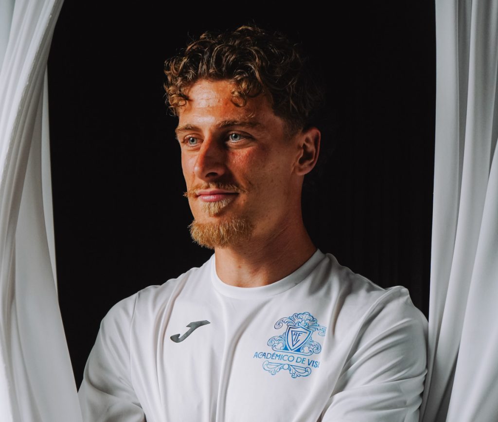 Portrait of a man with curly light brown hair and a short beard wearing a white athletic jersey, standing between white curtains against a dark background.