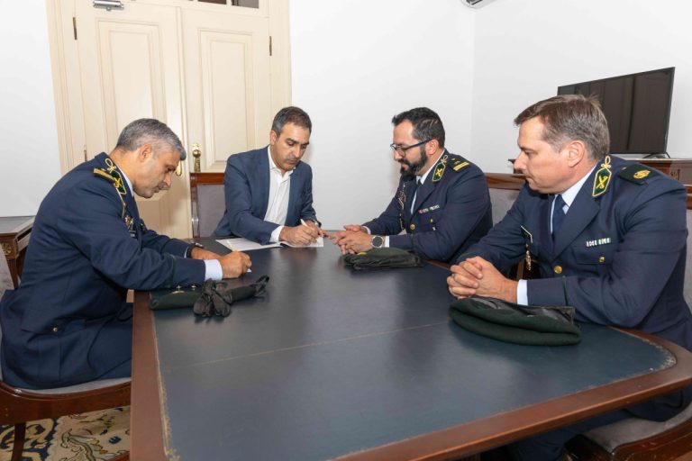 Four men sit around a large dark table as one in a suit signs a document while three in military uniforms look on in a formal office setting.
