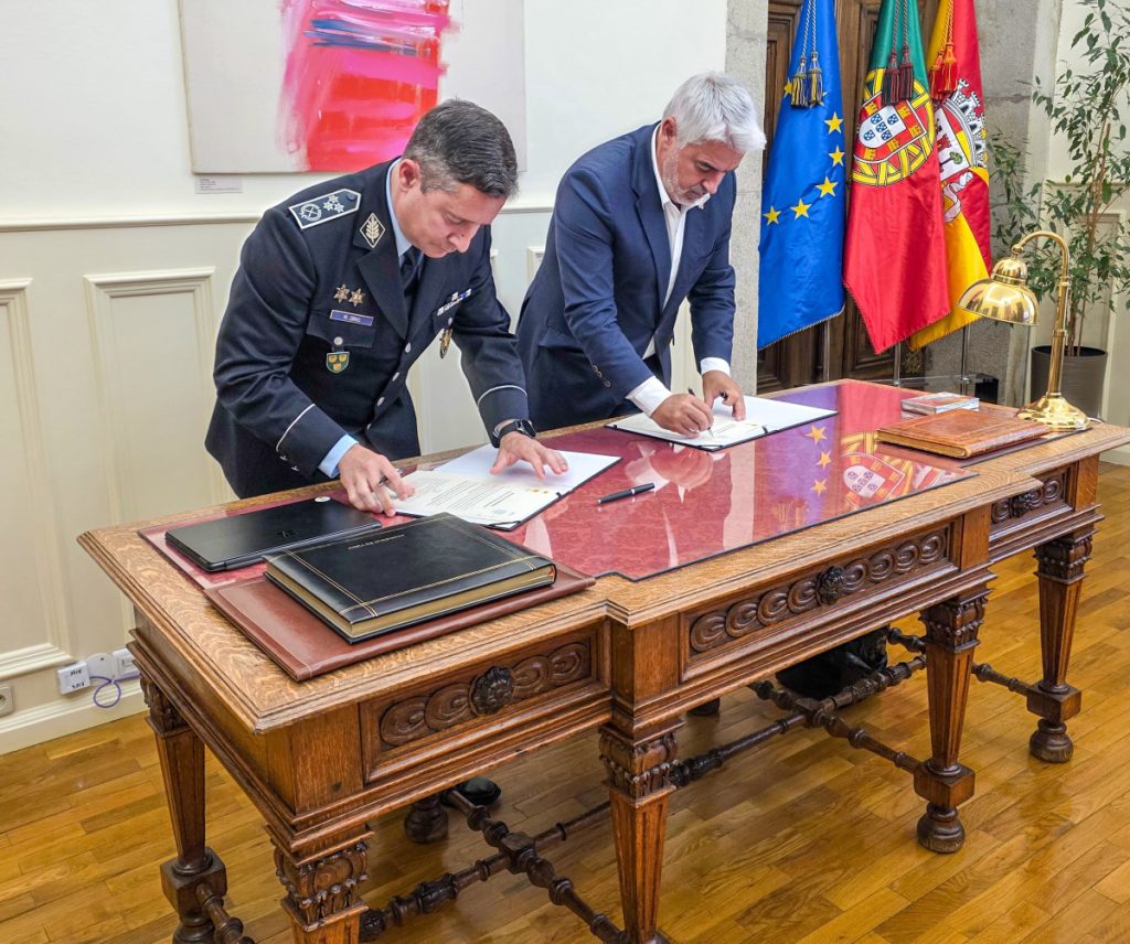 Two men sign documents on an ornate wooden desk in a formal office, with flags (EU and Spanish) behind them.