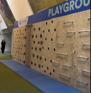 Indoor climbing gym wall with various handholds and holes, blue mats along the floor, and a person walking in the background under a 'PLAYGROUND' sign.