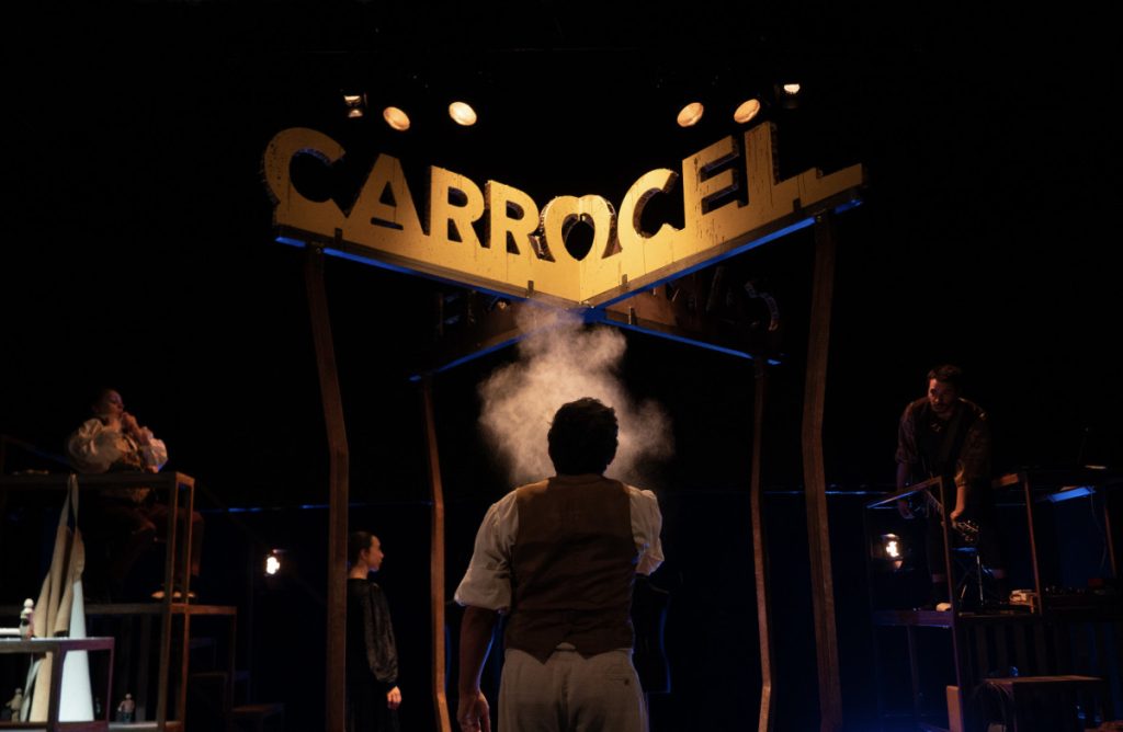 Person in vest stands center stage with steam billowing from behind, facing a lit sign that reads CARROCEL as a live scene unfolds with musicians on the right and a performer on a balcony to the left. ACERT celebra 50 anos em agosto com digressão do Trigo Limpo
