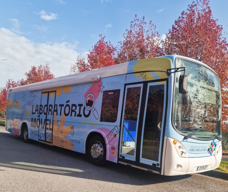 Colorful mobile laboratory bus with 'LABORATÓRIO MÓVEL' lettering parked on a street with red autumn trees in the background.