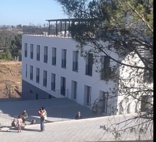 White multi-story building with tall vertical windows, a rooftop terrace, and people on a paved plaza in the foreground.