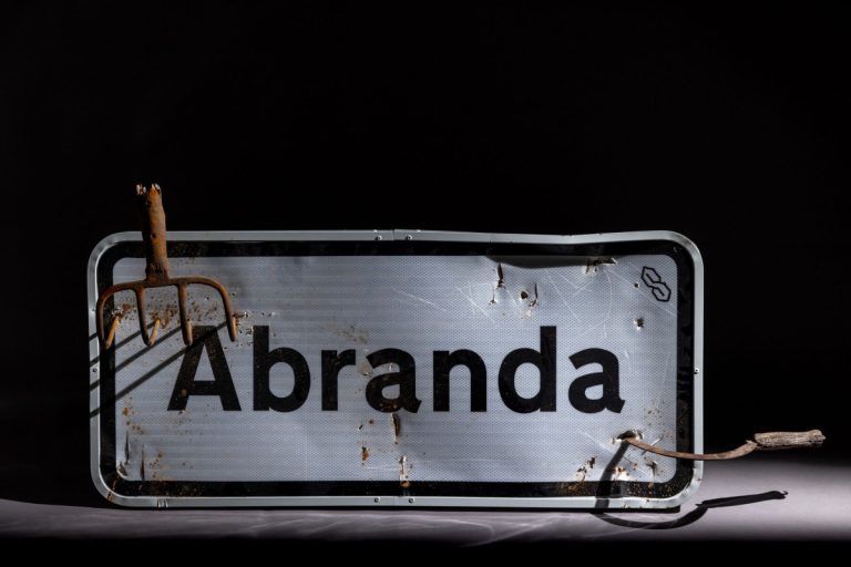 Old metal sign reading 'Abanda' with heavy rust and a rusted fork resting on the left, set against a dark background.