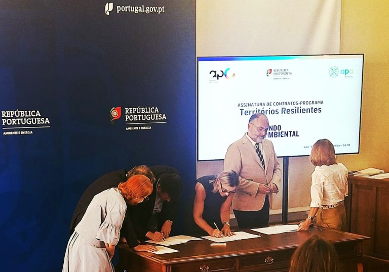 Officials signing documents at a formal signing ceremony, with a blue backdrop reading República Portuguesa and a large screen displaying 'Territórios Resilientes'.