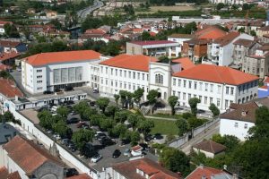Aerial view of a white university complex with red-tiled roofs, central grand entrance, and a tree-lined courtyard surrounded by parked cars and surrounding buildings.