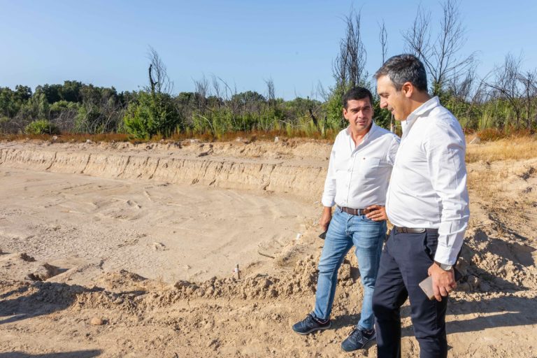Two men in white shirts stand at a dusty excavation site, examining the ground together.