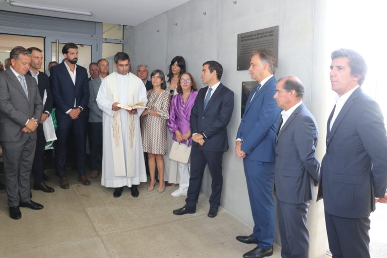 Group of formally dressed people listening as a priest reads from a book at an inauguration ceremony by a wall plaqueed with plaques.