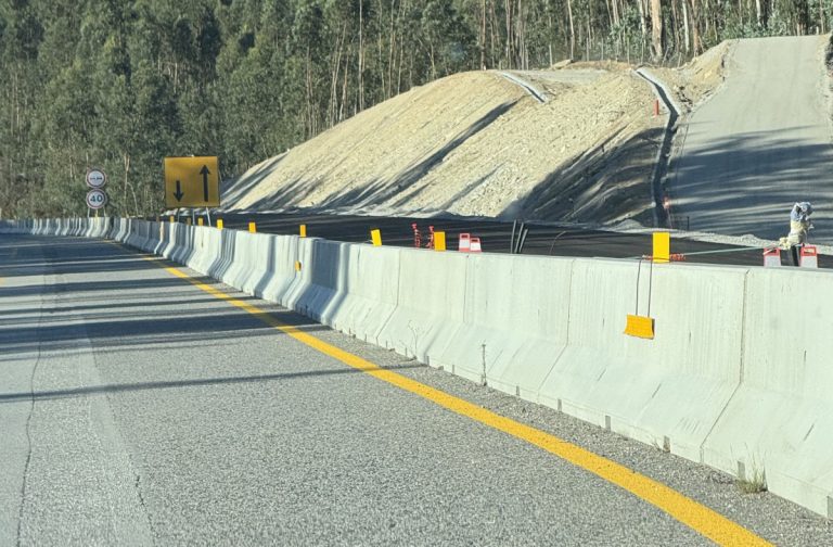 Highway under construction with concrete barriers, a dirt embankment, and yellow warning signs along the roadway edge at dusk/dawn light; no traffic on the open lane ahead.