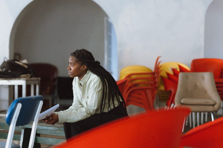 Woman with long braids sits on a bench, holding papers, in a room with stacked orange and yellow chairs in the background.