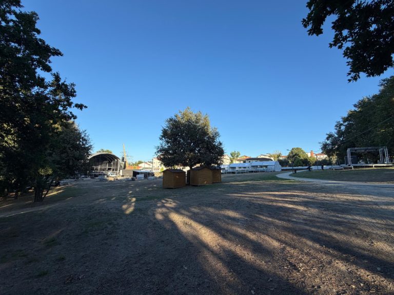 Open park area with a stage setup on the left, small wooden sheds grouped around a central tree, and white event tents in the background under a clear blue sky.