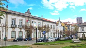 White municipal building with flags on a leafy street, cars parked along the curb, under a bright blue sky.