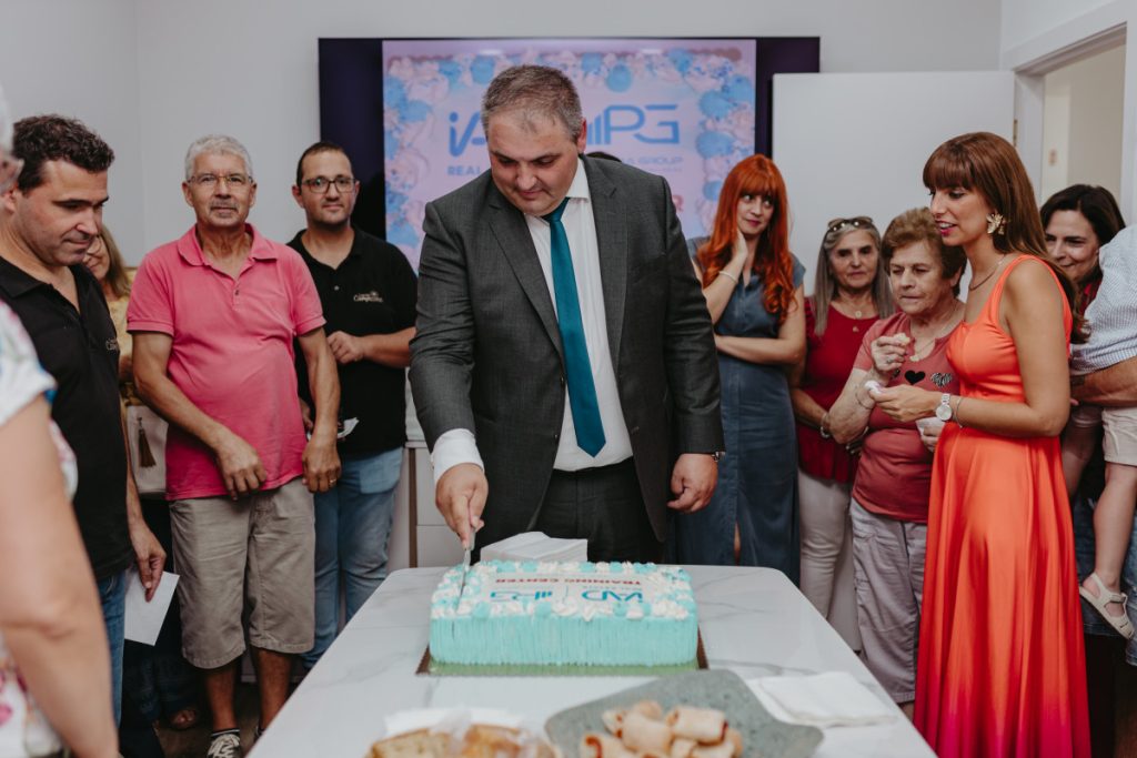 Man in a suit and teal tie cuts a rectangular cake as a group gathers around at a celebration in a bright room.