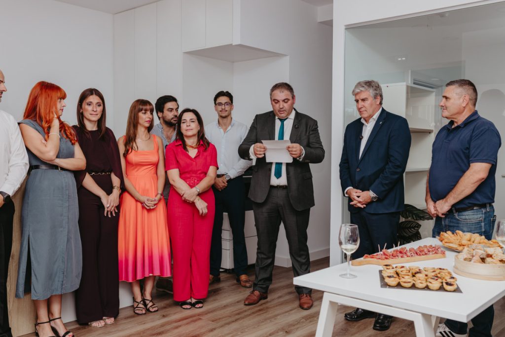 Group of professionals gathered in a bright kitchen as a man in a suit reads a speech to a small crowd with snacks on a table nearby.
