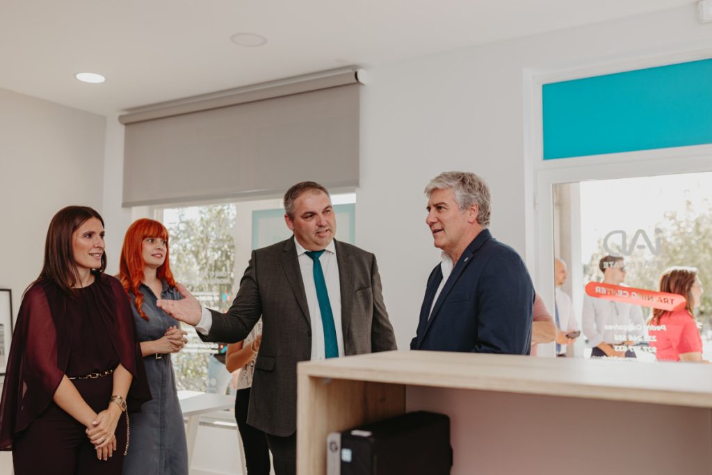 Four professionals in a bright office lobby chat; a man in a gray suit gestures toward a colleague in a navy blazer.