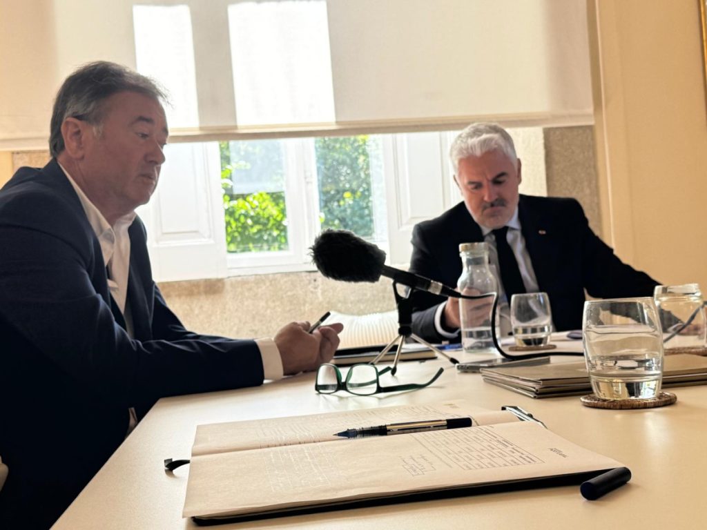 Two men in suits sit at a conference table with a microphone between them, reviewing documents and taking notes.