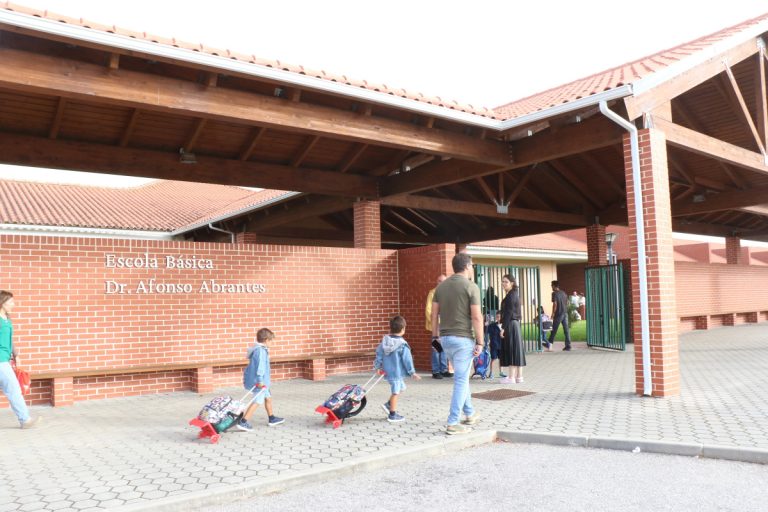 Children with rolling backpacks walk toward a brick school entrance under a wooden canopy; sign reads 'Escola Basica Dr. Afonso Abrantes'