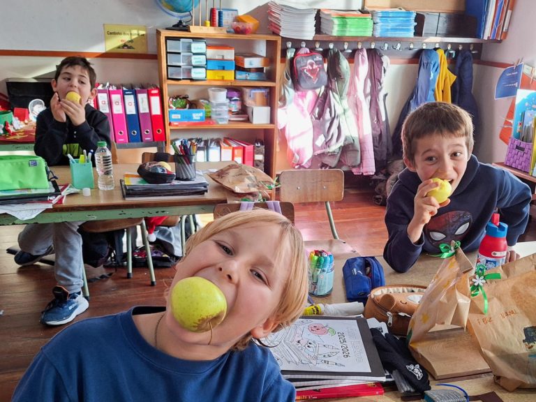 Three schoolchildren in a classroom eating apples at their desks, with backpacks, notebooks and coats hanging in the background.
