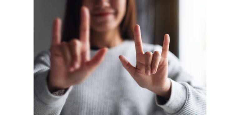 Person in a gray sweater makes ASL signs toward the camera, with both hands visible in the frame.