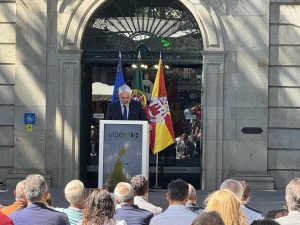 Man in a suit giving a speech at a podium outdoors, with flags behind him and an audience in front.