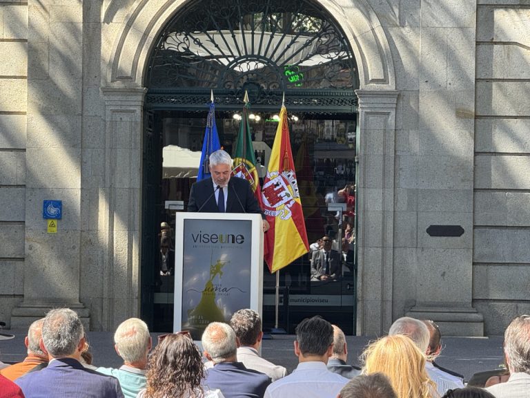 Man in a suit giving a speech at a podium outdoors, with flags behind him and an audience in front.