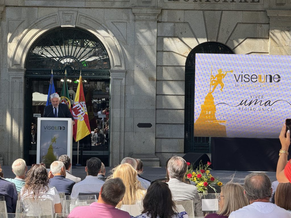 Man giving a speech at a podium during a formal outdoor event with flags in the background and a large screen banner to the right.