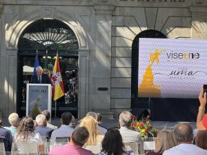 Man giving a speech at a podium during a formal outdoor event with flags in the background and a large screen banner to the right.