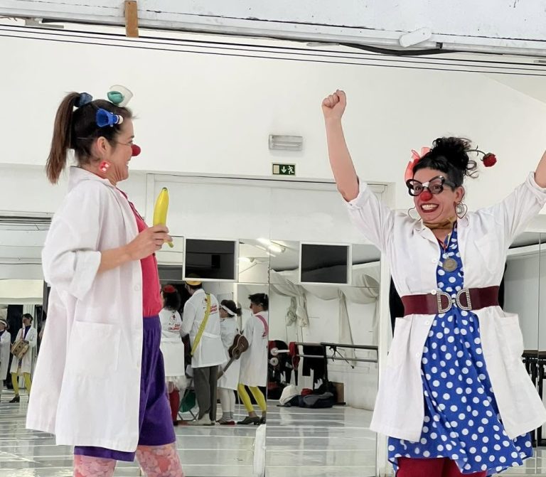 Two clowns in a rehearsal studio with mirrors, one holding a yellow banana-shaped prop while the other raises her arms in celebration.