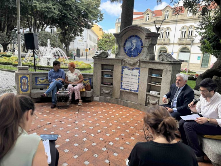 Public talk in a plaza: a microphone-wielding presenter speaks to a seated audience around a decorative stone monument.