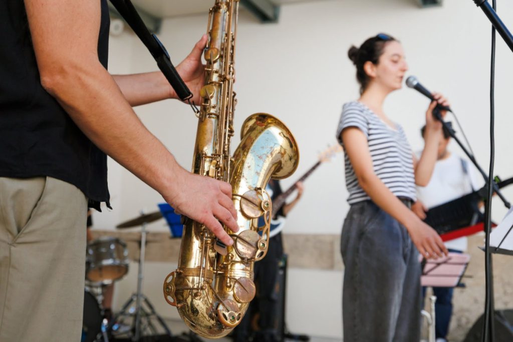 Close-up of a saxophonist holding a golden saxophone as a band performs in the background. The vocalist with a striped shirt sings into a microphone nearby.