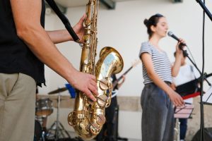 Close-up of a saxophonist holding a golden saxophone as a band performs in the background. The vocalist with a striped shirt sings into a microphone nearby.