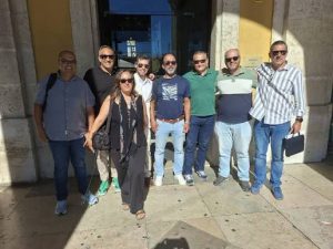 Nine adults posing together outside a building entrance on a sunny day, casually dressed.
