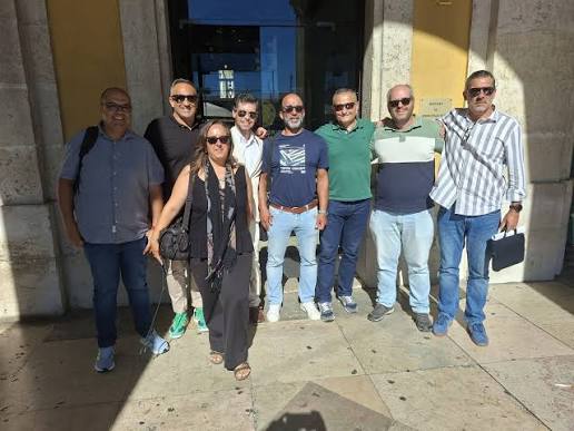 Nine adults posing together outside a building entrance on a sunny day, casually dressed.