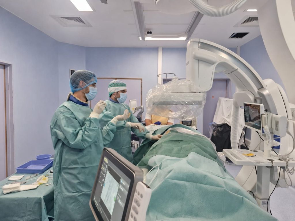 Surgeons in sterile gowns and masks perform surgery beside a patient on the operating table with imaging equipment overhead.