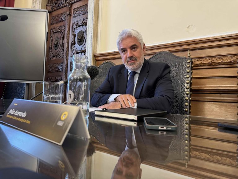 Man in a dark suit sits at a polished conference table with a microphone, water bottle, and notebook in front of him, in a formal meeting room.