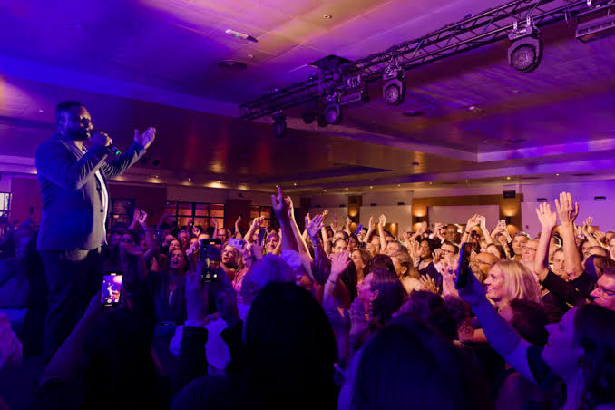 Male singer on stage performing to a packed crowd raising hands under purple lighting.