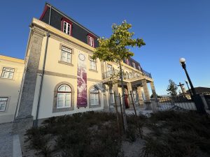 Historic beige building with stone accents, a second-floor balcony, and a purple banner, set against a clear blue sky; landscaped foreground with shrubs and a tree.