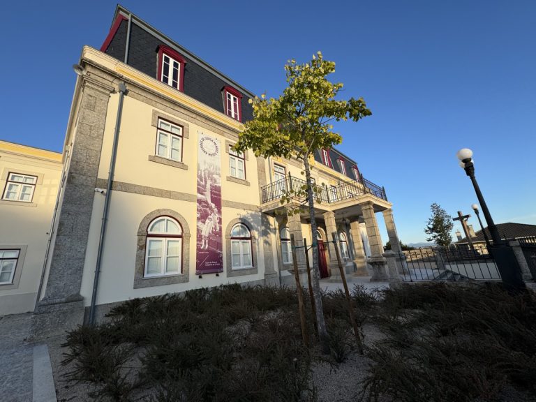 Historic beige building with stone accents, a second-floor balcony, and a purple banner, set against a clear blue sky; landscaped foreground with shrubs and a tree.