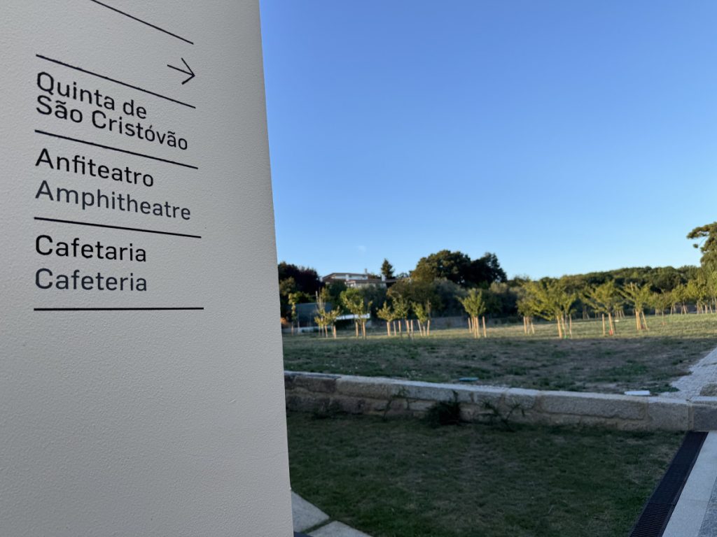 Vertical directional sign lists Quinta de São Cristóvão, Anfiteatro / Amphitheatre, Cafetaria / Cafeteria; park with young trees and a clear blue sky behind.