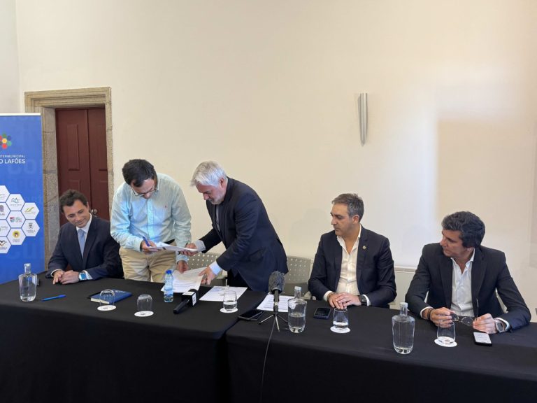 Five men in suits sit at a long table as two sign documents during a formal agreement ceremony, with microphones and water bottles on the table.